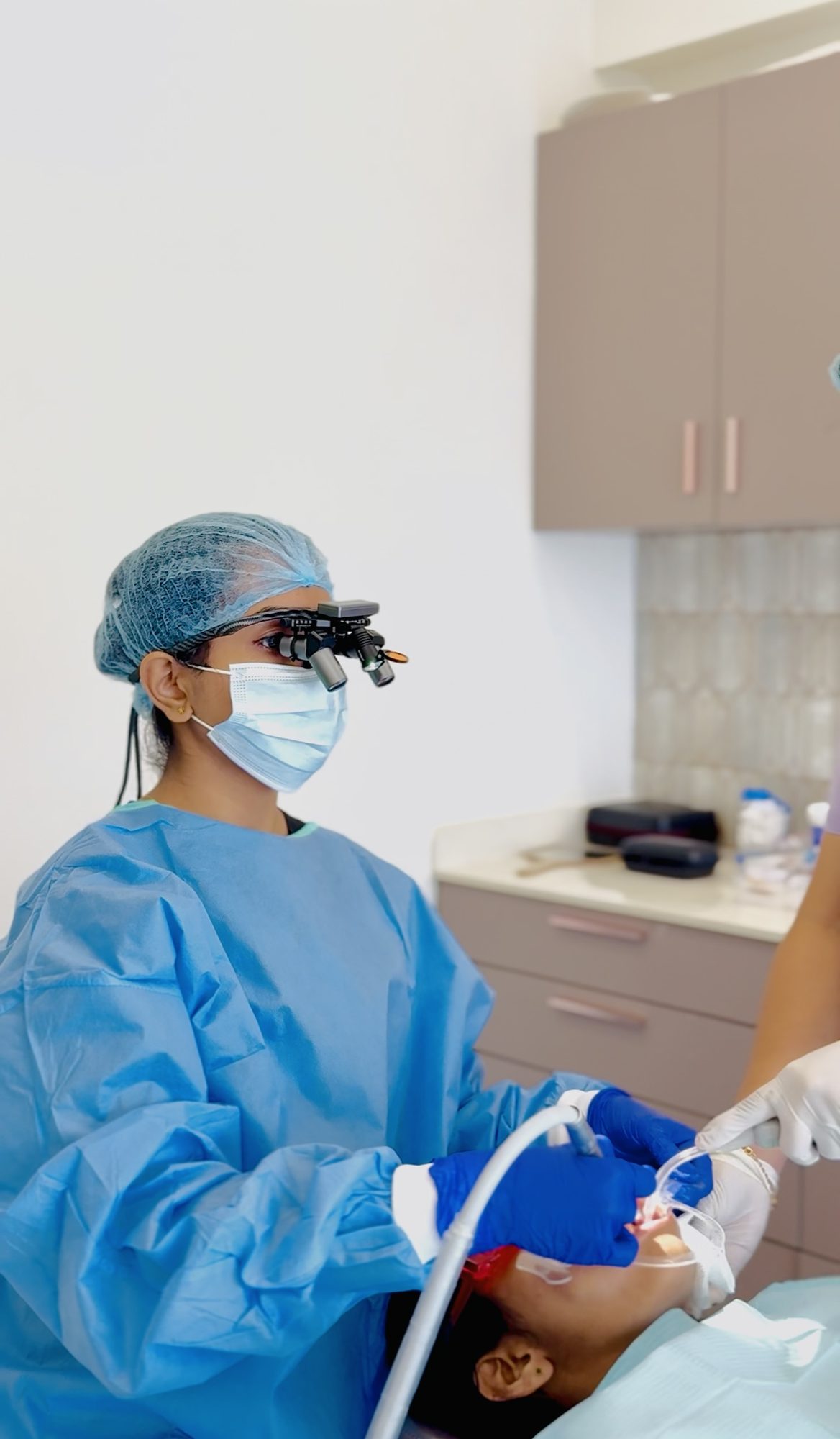 Dr. Alka Sajan treating a patient, wearing a surgical gown and magnifying loupes